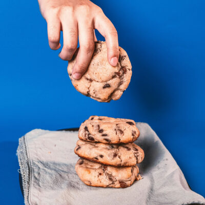 Eine Hand hebt vor blauem Hintergrund einen Schokokeks von einem Stapel hoch