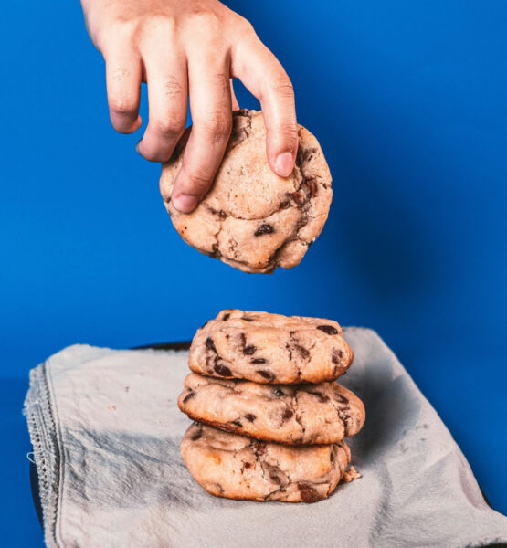 Eine Hand hebt vor blauem Hintergrund einen Schokokeks von einem Stapel hoch