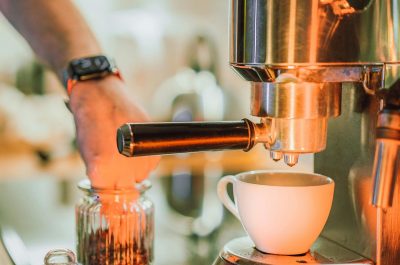 Hand bedient einen Siebträger an einer Edelstahl-Espressomaschine, davor eine weiße Tasse