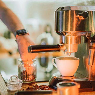 Hand bedient einen Siebträger an einer Edelstahl-Espressomaschine, davor eine weiße Tasse