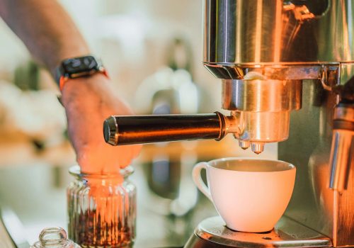 Hand bedient einen Siebträger an einer Edelstahl-Espressomaschine, davor eine weiße Tasse