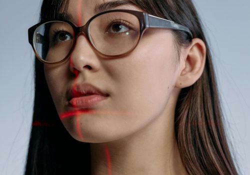 Eine Frau trägt eine Brille und schaut an der Kamera vorbei, während sich rote Laserstrahlen sich in ihrem Gesicht kreuzen