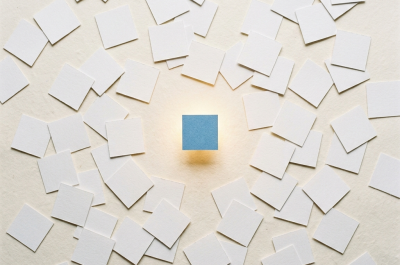 Dozens of identical small white squares are scattered randomly across a warm off-white surface — like physical index cards thrown on a desk. Each one casts a faint shadow. In the middle of the frame, one single Blue square glows faintly gold, slightly elevated above the rest, as if magnetically lifted. No text, no screens, no cables, no digital elements whatsoever. The mood is quiet, almost still-life. Shot from directly above, flat lay perspective, soft diffused daylight. Square format 1:1 aspect ratio, output as JPEG, file size under 1MB.