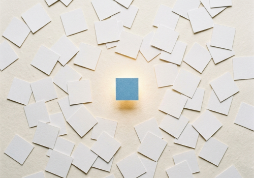 Dozens of identical small white squares are scattered randomly across a warm off-white surface — like physical index cards thrown on a desk. Each one casts a faint shadow. In the middle of the frame, one single Blue square glows faintly gold, slightly elevated above the rest, as if magnetically lifted. No text, no screens, no cables, no digital elements whatsoever. The mood is quiet, almost still-life. Shot from directly above, flat lay perspective, soft diffused daylight. Square format 1:1 aspect ratio, output as JPEG, file size under 1MB.