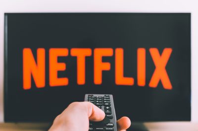 Im Hintergrund ist ein Fernseher mit dem Netflix-Logo, davor richtet eine Hand eine Fernbedienung darauf