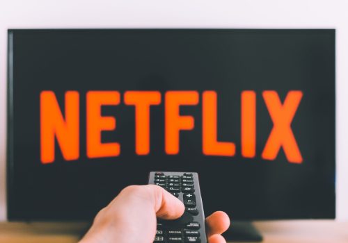 Im Hintergrund ist ein Fernseher mit dem Netflix-Logo, davor richtet eine Hand eine Fernbedienung darauf