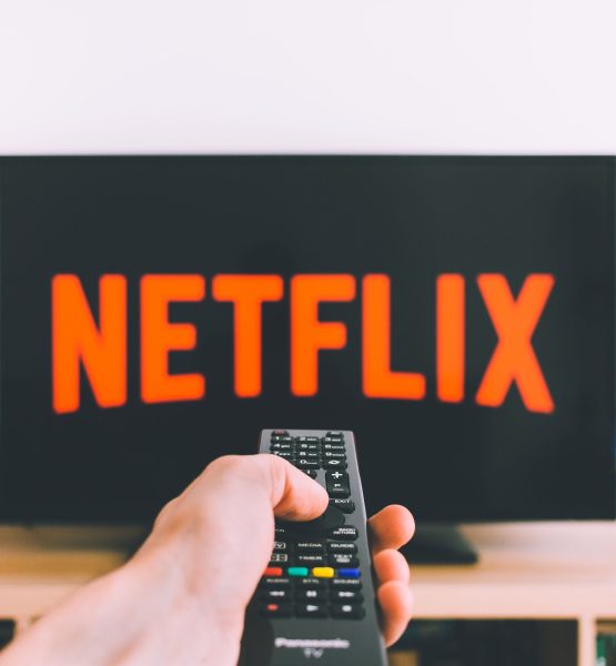 Im Hintergrund ist ein Fernseher mit dem Netflix-Logo, davor richtet eine Hand eine Fernbedienung darauf