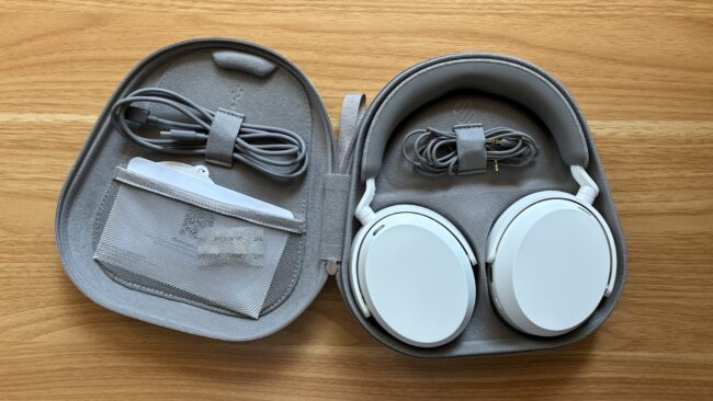 Geöffnete graue Sennheiser-Transporttasche auf Holzuntergrund. In der oberen Hälfte ein weißer Over-Ear-Kopfhörer sowie ein aufgewickeltes Klinkenkabel. In der unteren Hälfte ein USB-C-Ladekabel und eine transparente Netztasche mit Dokumenten.