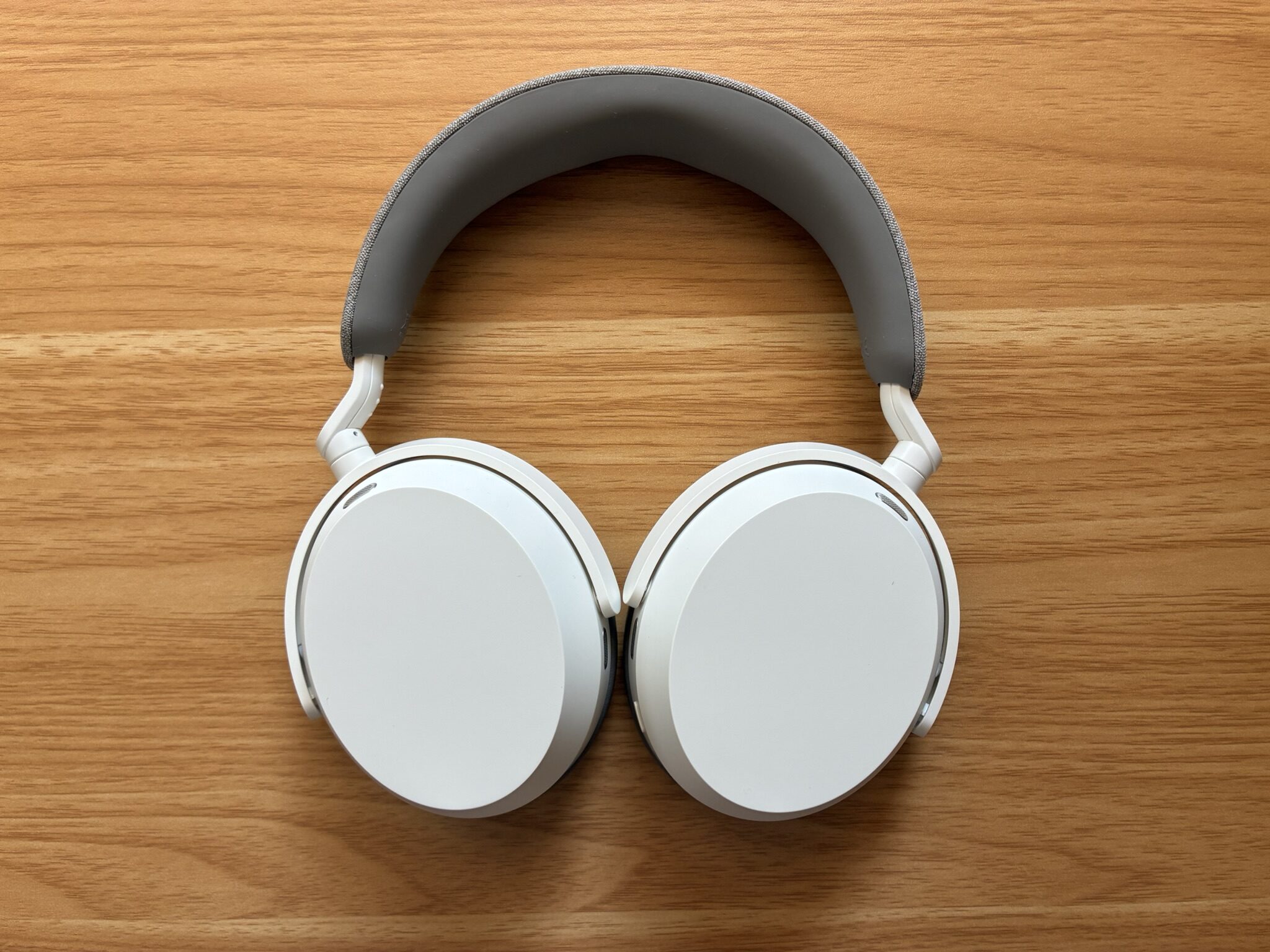 Weißer Over-Ear-Kopfhörer von oben auf hellem Holzuntergrund. Grau gepolstertes Kopfband, weiße Ohrmuscheln mit glatter Außenfläche und silberfarbenen Scharnieren. Klares, minimalistisches Design.