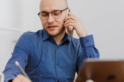 Eine Person mit Glatze und schwarzer Brille sitzt an einem Schreibtisch, telefoniert mit einem Smartphone und macht gleichzeitig Notizen in einem Notizbuch. Sie trägt ein blaues Hemd mit weißem Punktmuster. Auf dem Tisch stehen ein Tablet auf einem Ständer und eine weiße Tasse. Heller, unscharfer Bürohintergrund.