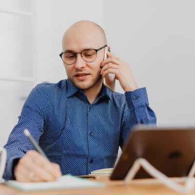 Eine Person mit Glatze und schwarzer Brille sitzt an einem Schreibtisch, telefoniert mit einem Smartphone und macht gleichzeitig Notizen in einem Notizbuch. Sie trägt ein blaues Hemd mit weißem Punktmuster. Auf dem Tisch stehen ein Tablet auf einem Ständer und eine weiße Tasse. Heller, unscharfer Bürohintergrund.