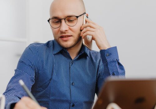 Eine Person mit Glatze und schwarzer Brille sitzt an einem Schreibtisch, telefoniert mit einem Smartphone und macht gleichzeitig Notizen in einem Notizbuch. Sie trägt ein blaues Hemd mit weißem Punktmuster. Auf dem Tisch stehen ein Tablet auf einem Ständer und eine weiße Tasse. Heller, unscharfer Bürohintergrund.