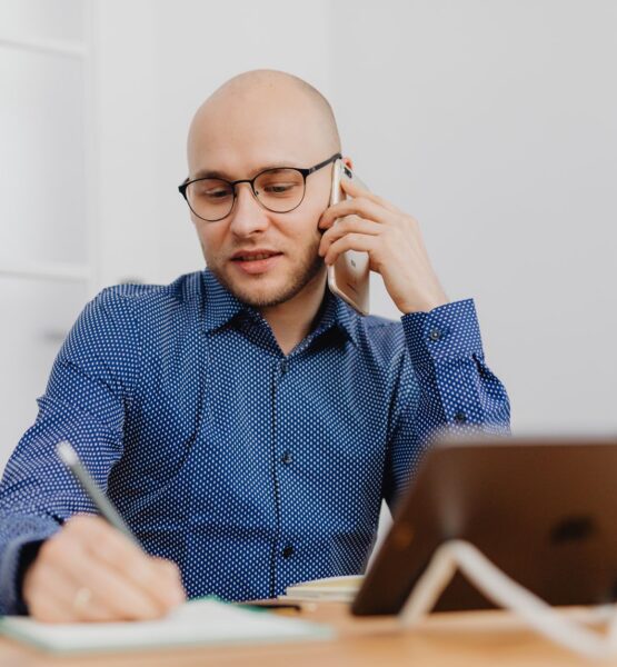 Eine Person mit Glatze und schwarzer Brille sitzt an einem Schreibtisch, telefoniert mit einem Smartphone und macht gleichzeitig Notizen in einem Notizbuch. Sie trägt ein blaues Hemd mit weißem Punktmuster. Auf dem Tisch stehen ein Tablet auf einem Ständer und eine weiße Tasse. Heller, unscharfer Bürohintergrund.
