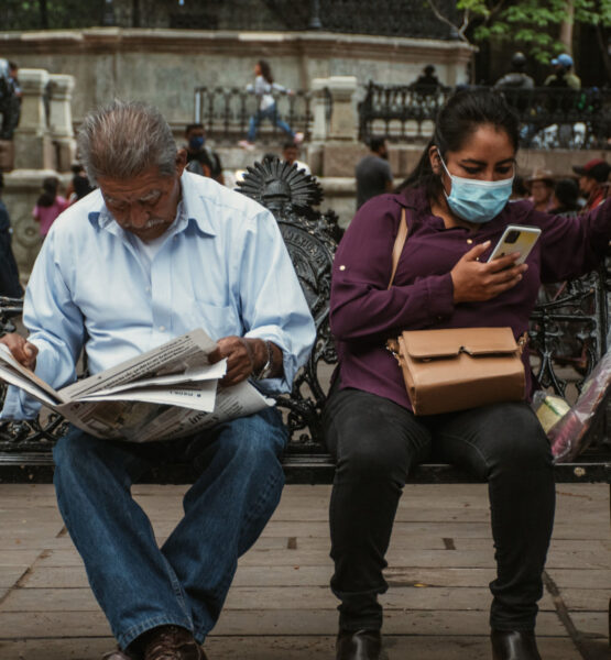 Zwei Personen sitzen nebeneinander auf einer Bank, der Mann links liest eine Zeitung, die junge Frau rechts schaut auf ihr Smartphone