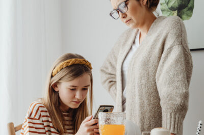 Ein junges Mädchen hängt am Frühstückstisch vor ihrem Handy während ihre Mutter genervt daneben steht.
