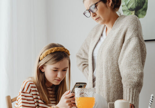 Ein junges Mädchen hängt am Frühstückstisch vor ihrem Handy während ihre Mutter genervt daneben steht.
